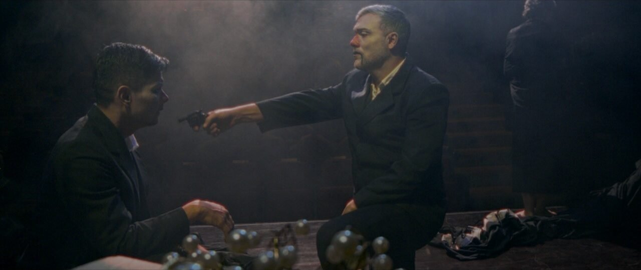 Escena de suspenso en un escenario oscuro y con humo, donde un hombre de pie con traje y maquillaje teatral apunta un revólver a otro hombre sentado frente a él. Fotograma del cortometraje El Telon de los Grillos