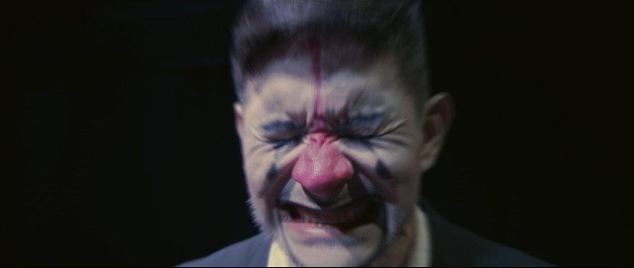 Primer plano del rostro de un actor con maquillaje de payaso desalineado, cerrando los ojos con fuerza y apretando los dientes en una clara expresión de dolor o llanto.