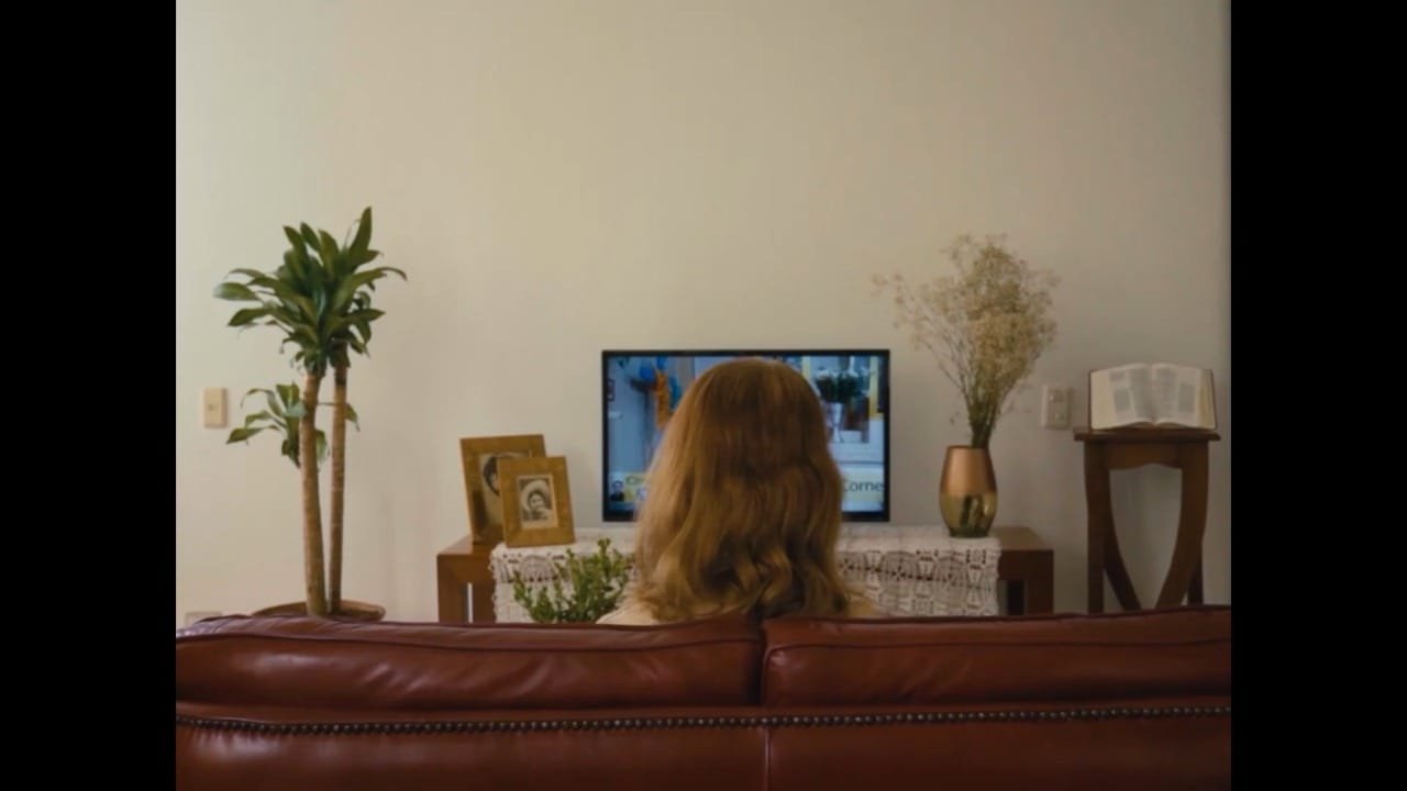 Toma de espalda de una mujer pelirroja sentada en un sofá de cuero marrón viendo la televisión en una sala de estar decorada con plantas y portarretratos