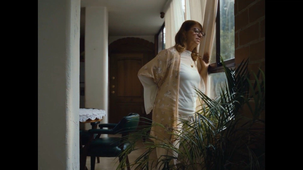 Mujer madura con gafas y un kimono beige de pie junto a una gran ventana mirando hacia afuera con expresión pensativa, en un interior con plantas y muebles oscuros.