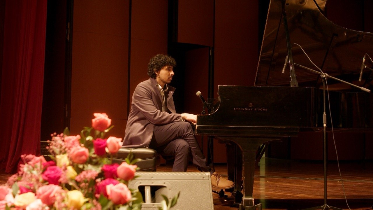Hombre vestido de traje tocando el piano en un escenario, con flores adornando