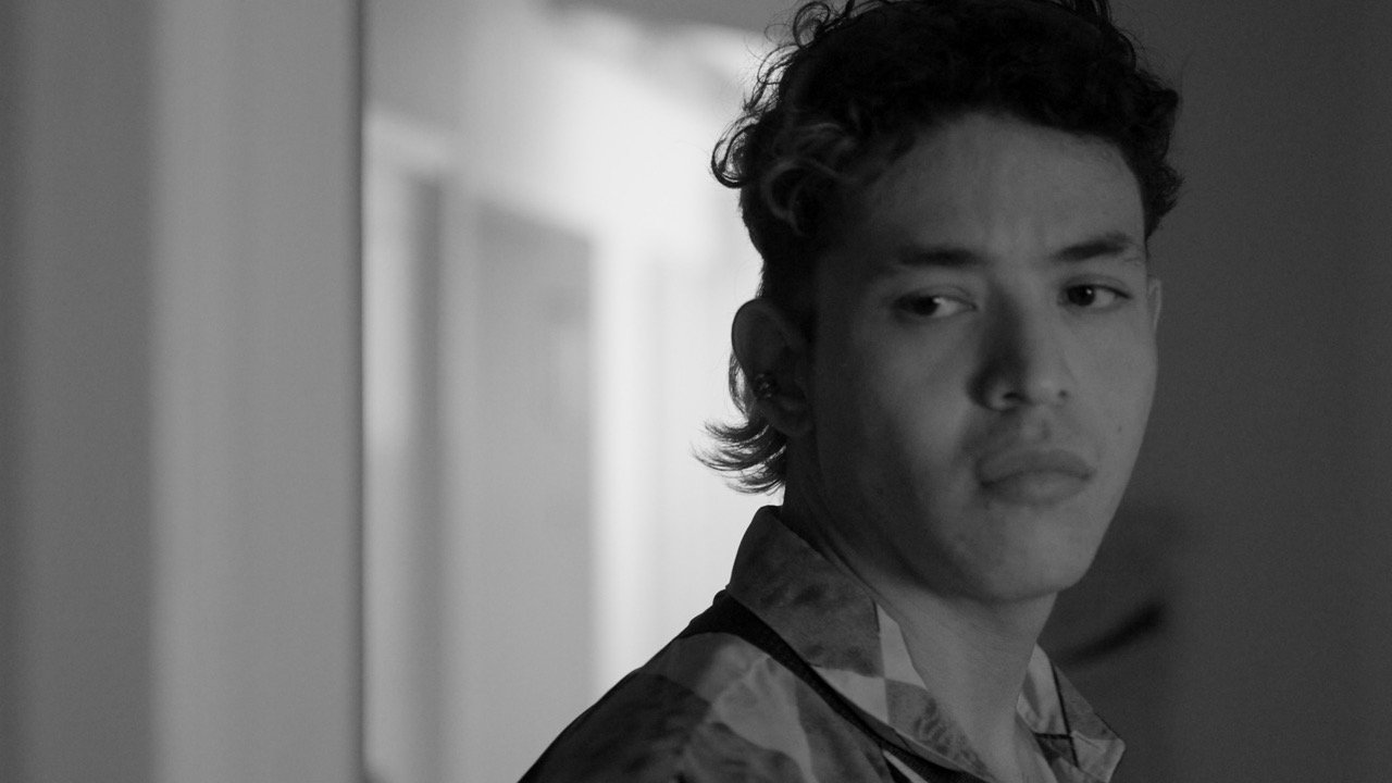 Fotograma en blanco y negro de un joven con cabello rizado y camisa estampada mirando pensativo sobre su hombro derecho hacia un pasillo iluminado