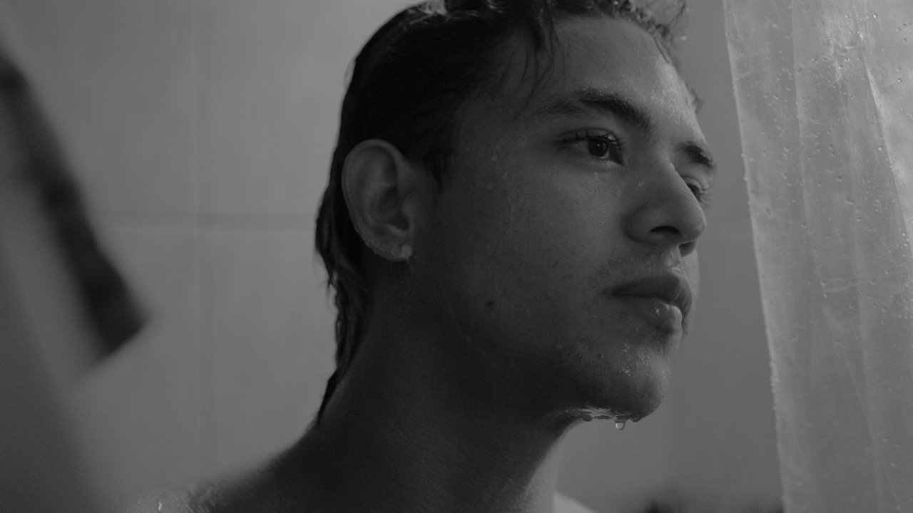 Toma cinematográfica en blanco y negro de perfil de un joven con el rostro y cabello mojados dentro de una ducha, mirando reflexivamente a través de una cortina translúcida.