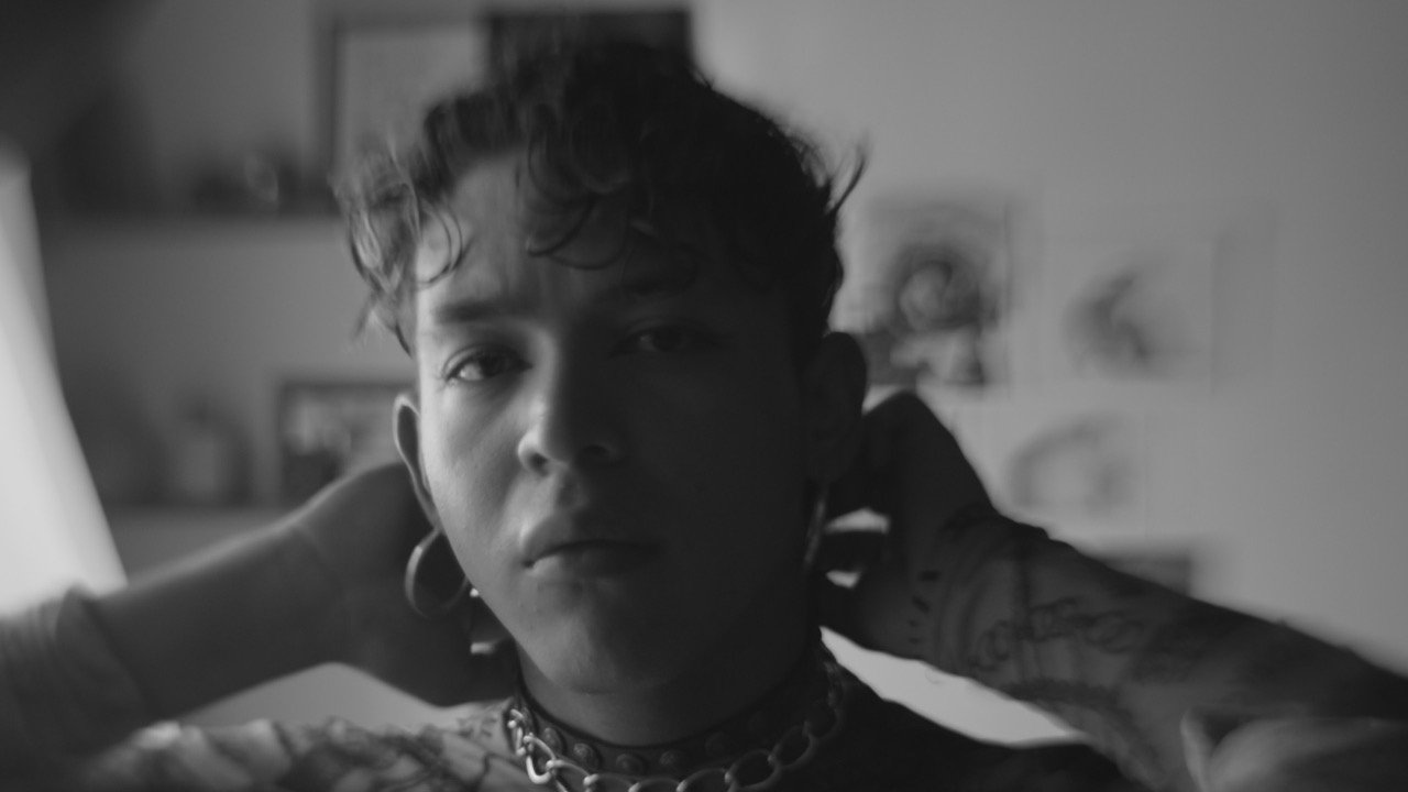 Retrato en blanco y negro de un joven con expansores en las orejas, mirando fijamente a la cámara mientras se ajusta un collar de cadena gruesa en el cuello, con fotografías desenfocadas en el fondo
