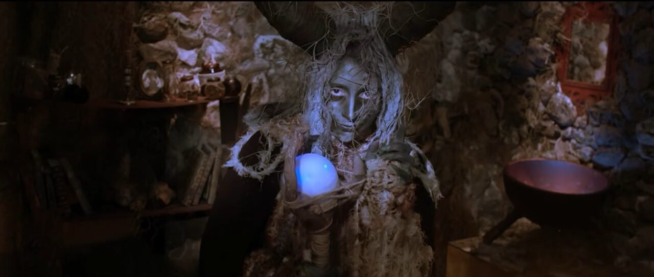 Primer plano de una persona mimetizada con madera seca y maleza retroiluminado por una luz azul brillante, simulando un fuego mágico o una entrada misteriosa