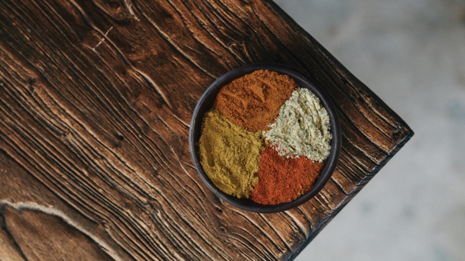 Cuenco con cuatro tipos de especias molidas en colores amarillo, naranja, blanco y rojo, ubicado sobre una tabla rústica de madera oscura.