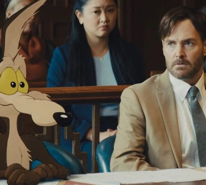 Escena de tribunal híbrida de acción real y animación de la película Coyote vs. Acme, mostrando al personaje animado Wile E. Coyote sentado en la mesa de la defensa junto al actor Will Forte, quien interpreta a su abogado, con el jurado al fondo