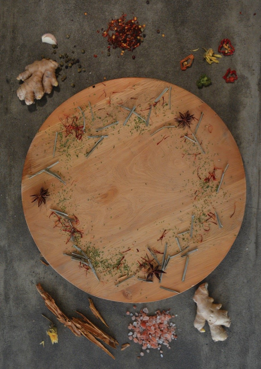 Vista superior de una tabla de cortar de madera circular rodeada de ingredientes enteros como bulbos de jengibre fresco, anís estrellado, copos de pimiento rojo y sal marina rosada sobre una superficie gris texturizada
