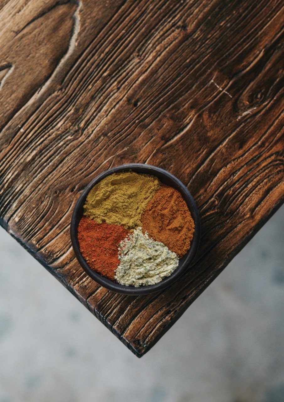 Cuenco con cuatro tipos de especias molidas en colores amarillo, naranja, blanco y rojo, ubicado sobre una tabla rústica de madera oscura.