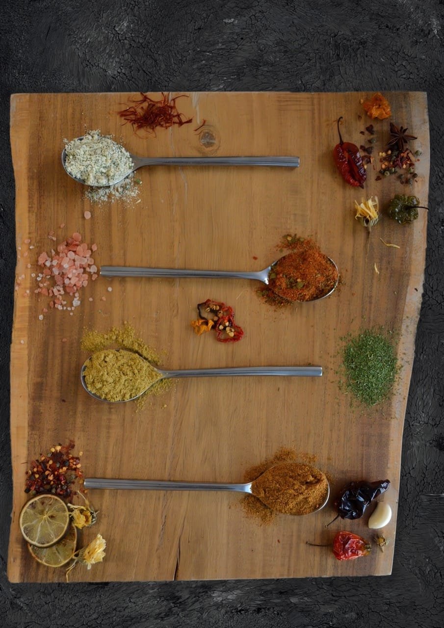 Vista aérea de una tabla de madera rústica de borde vivo con cuatro cucharas de metal llenas de diferentes especias, rodeadas de azafrán, anís estrellado y copos de pimiento rojo