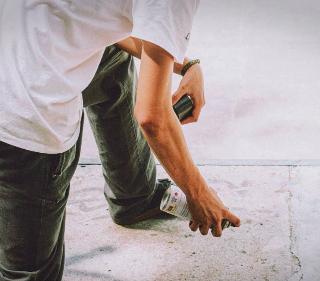 Fotografía de cerca que detalla a una persona con camiseta blanca y pantalones oscuros sosteniendo una lata de pintura en aerosol blanca a punto de marcar el piso de cemento
